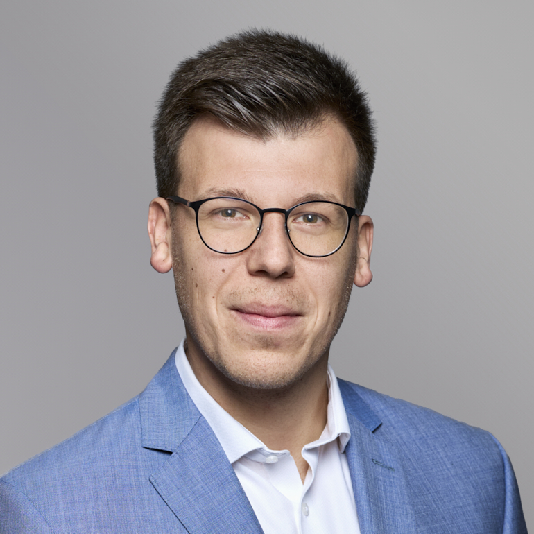 Ein junger Mann mit Brille und einem hellblauen Anzugsmantel lächelt leicht. Er hat kurzes, dunkles Haar und steht vor einem grauen Hintergrund.