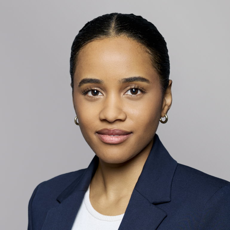 Ein Porträt einer jungen Frau mit dunkelblonden, glatten Haaren, die zu einem Zopf gebunden sind. Sie trägt ein dunkles Jackett über einem weißen Oberteil und schaut direkt in die Kamera. Ihr Gesichtsausdruck ist freundlich und selbstbewusst. Der Hintergrund ist einfarbig und neutral.