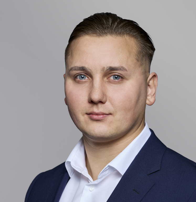 Ein junger Mann mit kurzen, welligen Haaren und blauen Augen trägt ein dunkelblaues Sakko und ein weißes Hemd. Er schaut direkt in die Kamera, mit einem neutralen Gesichtsausdruck vor einem hellen Hintergrund.