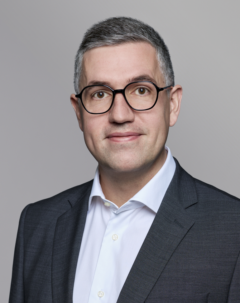 Porträt eines Mannes mit grauen Haaren und Brille, der in einem dunkelgrauen Anzug und einem weißen Hemd gekleidet ist. Er blickt direkt in die Kamera und wirkt freundlich und professionell. Der Hintergrund ist neutral und trägt zur Fokussierung auf seine Person bei.