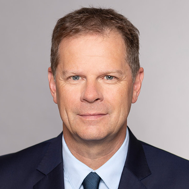A professional headshot of a middle-aged man with short brown hair, wearing a dark suit and a light blue shirt with a tie. He is looking directly at the camera with a neutral expression against a plain light gray background.