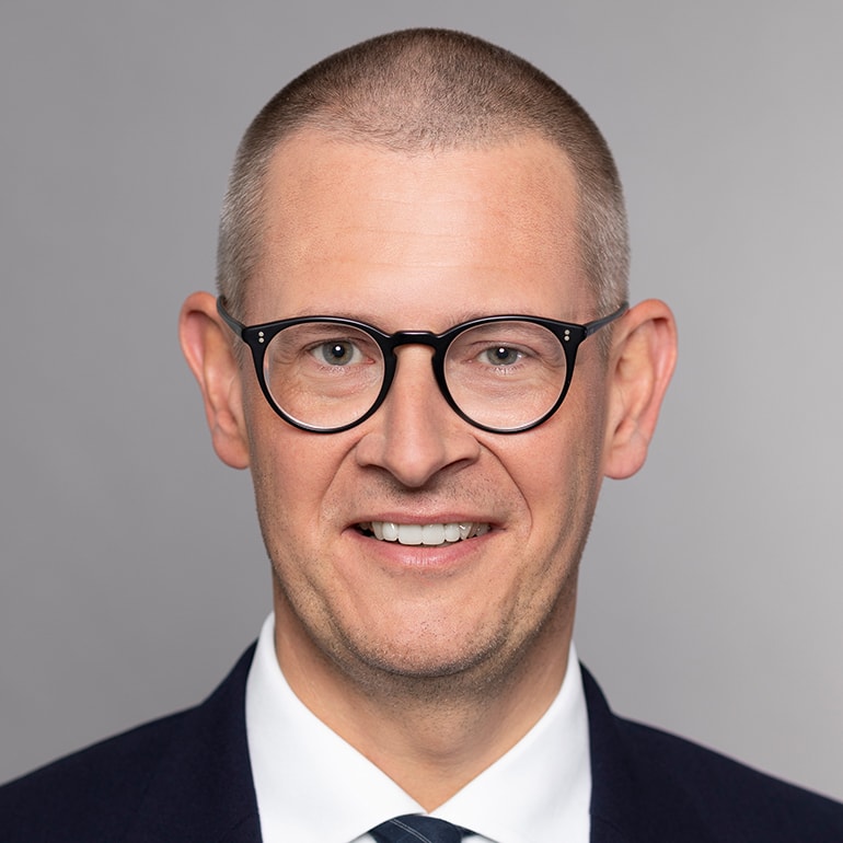 Portrait of a man with a shaved head and glasses, smiling directly at the camera. He wears a dark suit and a white shirt with a tie. The background is a neutral gray, emphasizing his friendly expression.