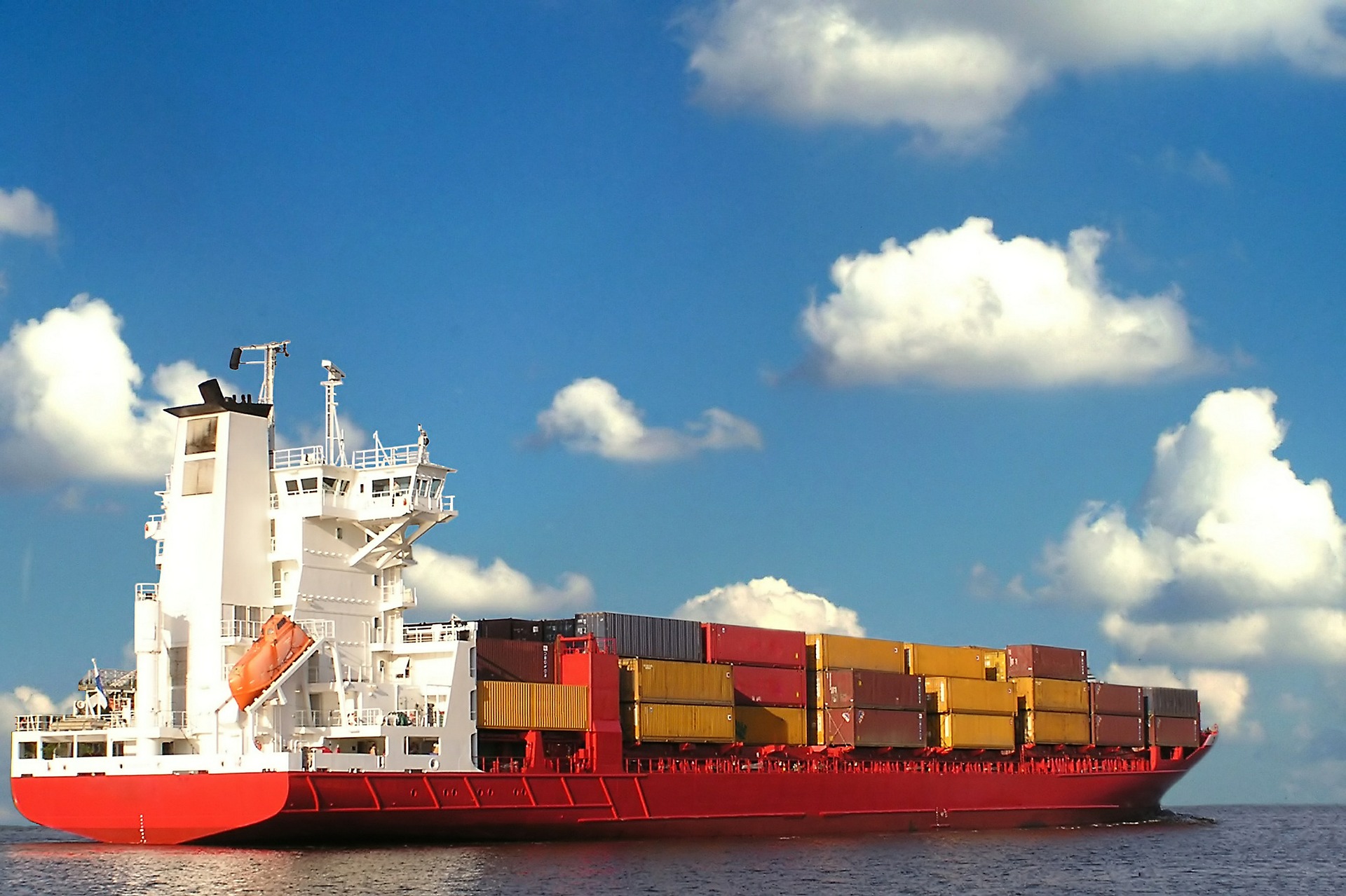 A large container ship with a red hull is anchored on calm waters, displaying numerous colorful shipping containers stacked on its deck. The sky above features a few fluffy white clouds, creating a bright and clear backdrop.