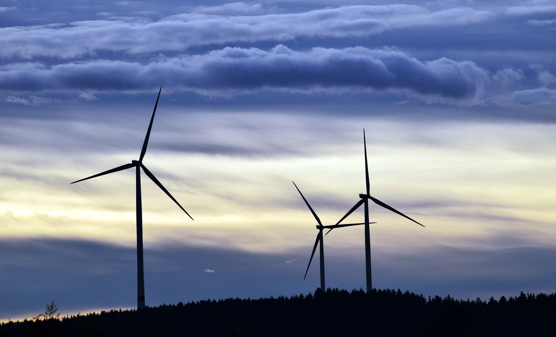 Three wind turbines stand silhouetted against a twilight sky, with gentle clouds illuminated by soft, fading light. The turbines represent renewable energy and sustainability, standing amidst a landscape of trees.
