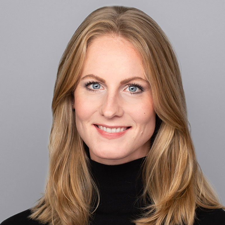 A woman with long, light brown hair is smiling directly at the camera. She is wearing a black turtleneck and has blue eyes. The background is a neutral gray, enhancing her friendly expression.