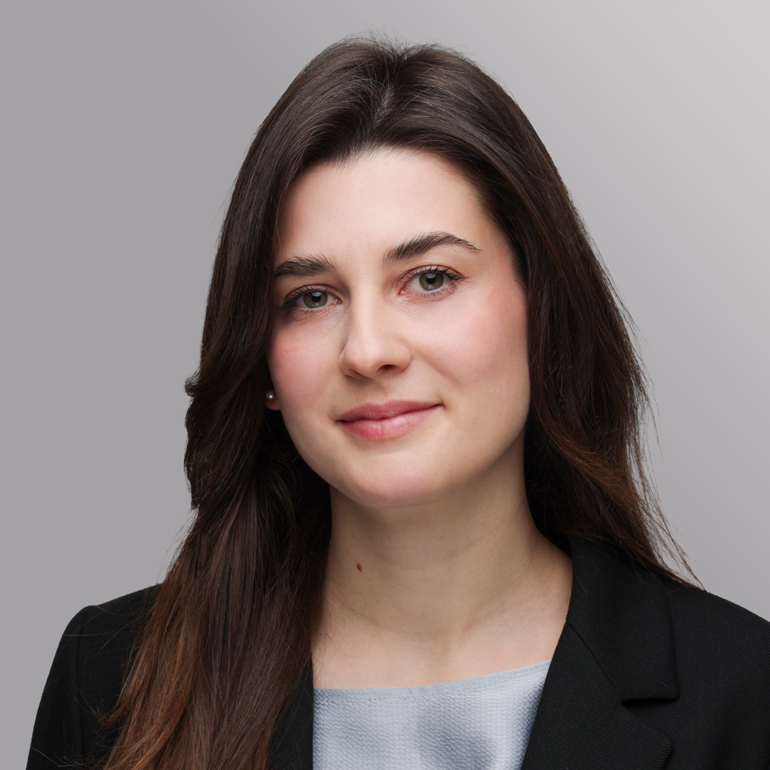 Ein Porträt einer jungen Frau mit langen, braunen Haaren. Sie trägt einen schwarzen Blazer und hat ein sanftes Lächeln. Der Hintergrund ist neutral und hell, wodurch ihr Gesicht gut zur Geltung kommt.