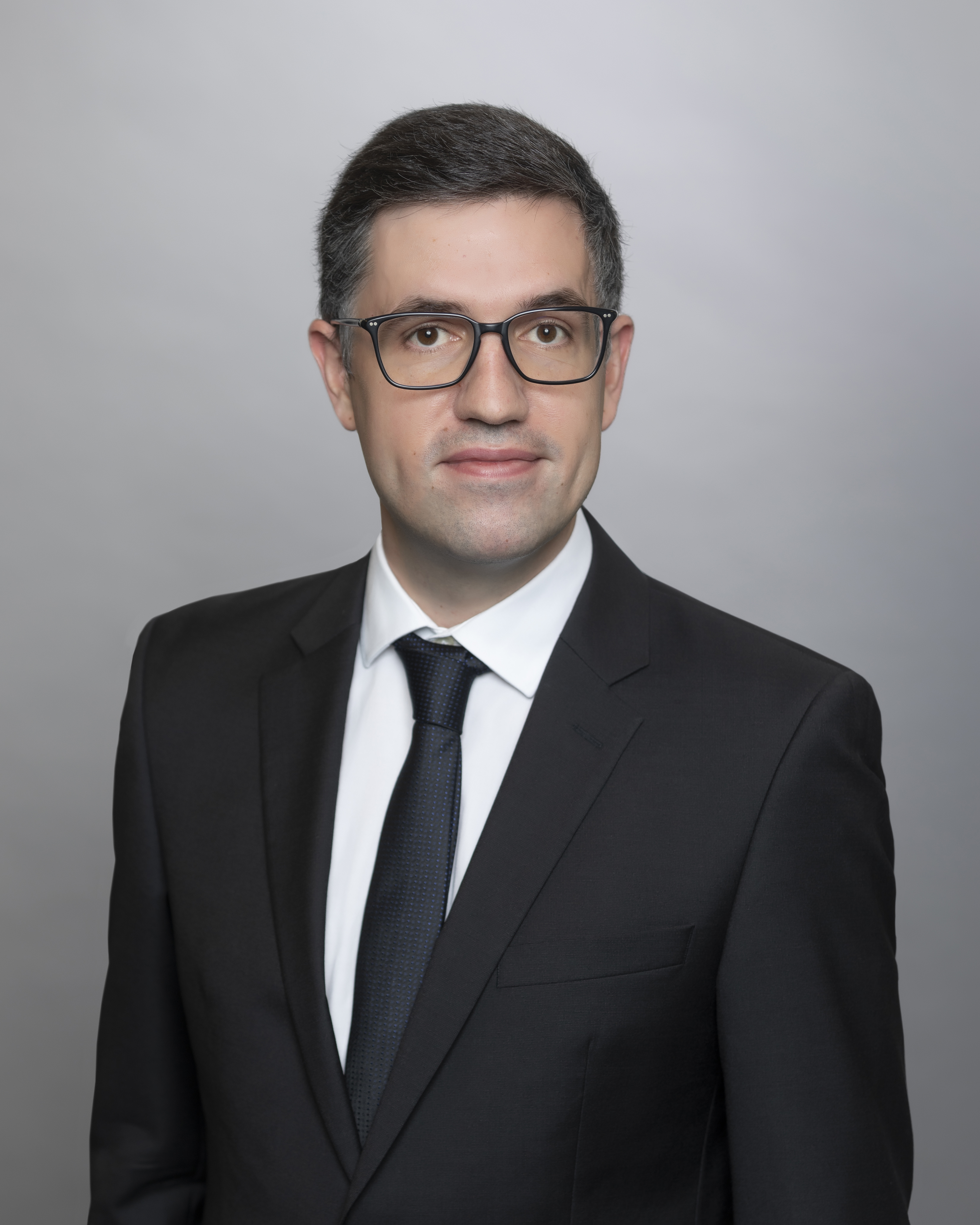 A professional man wearing glasses and a black suit poses against a light gray background. He has short, dark hair and a neutral expression, exuding confidence and professionalism.