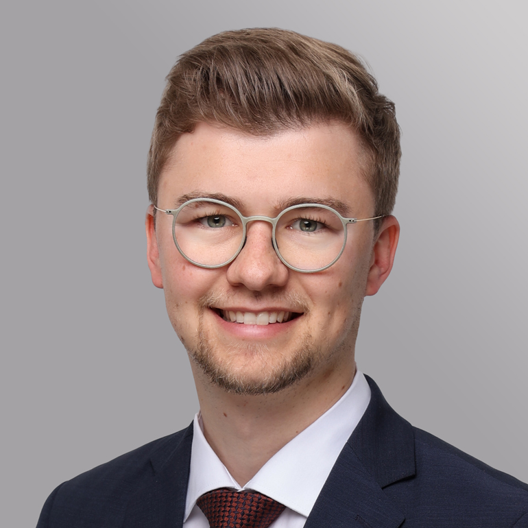 A young man with short, light brown hair and glasses smiles in a formal suit and tie. The background is a soft gray gradient, emphasizing his cheerful expression. He appears professional and approachable.