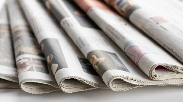 Auf dem Bild sind mehrere gefaltete Zeitungen zu sehen, die übereinander liegen. Die Zeitungen sind in neutralen Farben gehalten und zeigen einige Bilder samt Text auf der Vorderseite. Sie vermitteln den Eindruck von Vielfalt in den Themen.