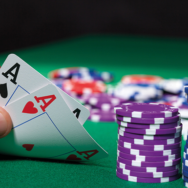 A hand holding two Ace of hearts and Ace of spades is placed near a stack of purple and white poker chips on a green felt surface, suggesting an ongoing card game.