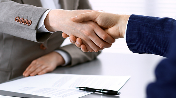Händedruck zwischen zwei Personen, die dabei an einem Tisch sitzen. Auf dem Tisch liegen Dokumente und ein Stift. Die Personen scheinen eine Vereinbarung oder einen Vertrag abzuschließen.