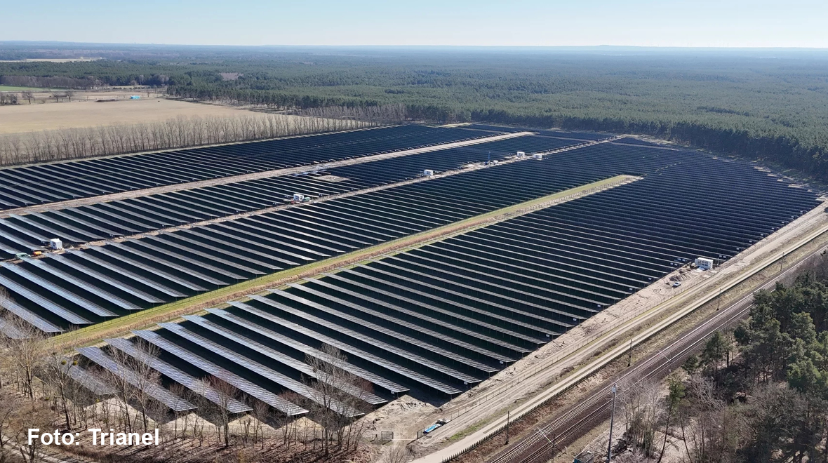 Luftaufnahme einer großen Solaranlage, die sich über mehrere Hektar erstreckt. Die Fläche ist mit zahlreichen solaren Paneelen bedeckt, die in langen Reihen angeordnet sind. Im Hintergrund sind Wälder und ein Bahnübergang sichtbar.