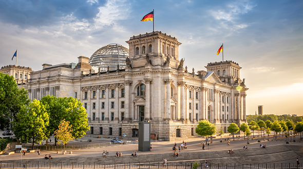 The image shows the Reichstag building in Berlin, featuring its ornate architecture and glass dome. Flags flutter from the roof, and people walk in the surrounding area, which includes greenery and pathways. The scene is illuminated by warm sunset light.