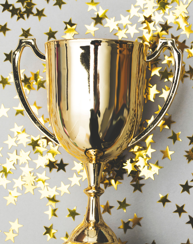 A shiny gold trophy stands upright against a gray background, surrounded by scattered golden star-shaped confetti. The trophy features two handles and a smooth, reflective surface.