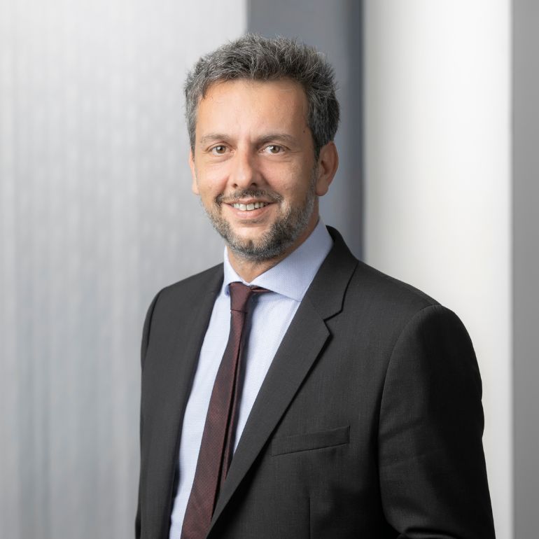 A man with short, greying hair and a beard smiles while wearing a black suit, white shirt, and maroon tie. The background features a light, textured wall, enhancing the professional atmosphere of the portrait.