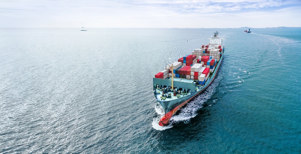 A large container ship sails through calm ocean waters, prominently displaying colorful shipping containers on its deck. The vessel creates a wake as it moves, with a distant horizon and a few other boats visible in the background.