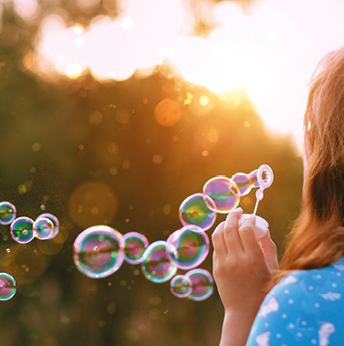 A child with long hair is blowing bubbles into the air during sunset. The bubbles are large and colorful, floating upwards against a golden and blurred background, creating a joyful and whimsical atmosphere.