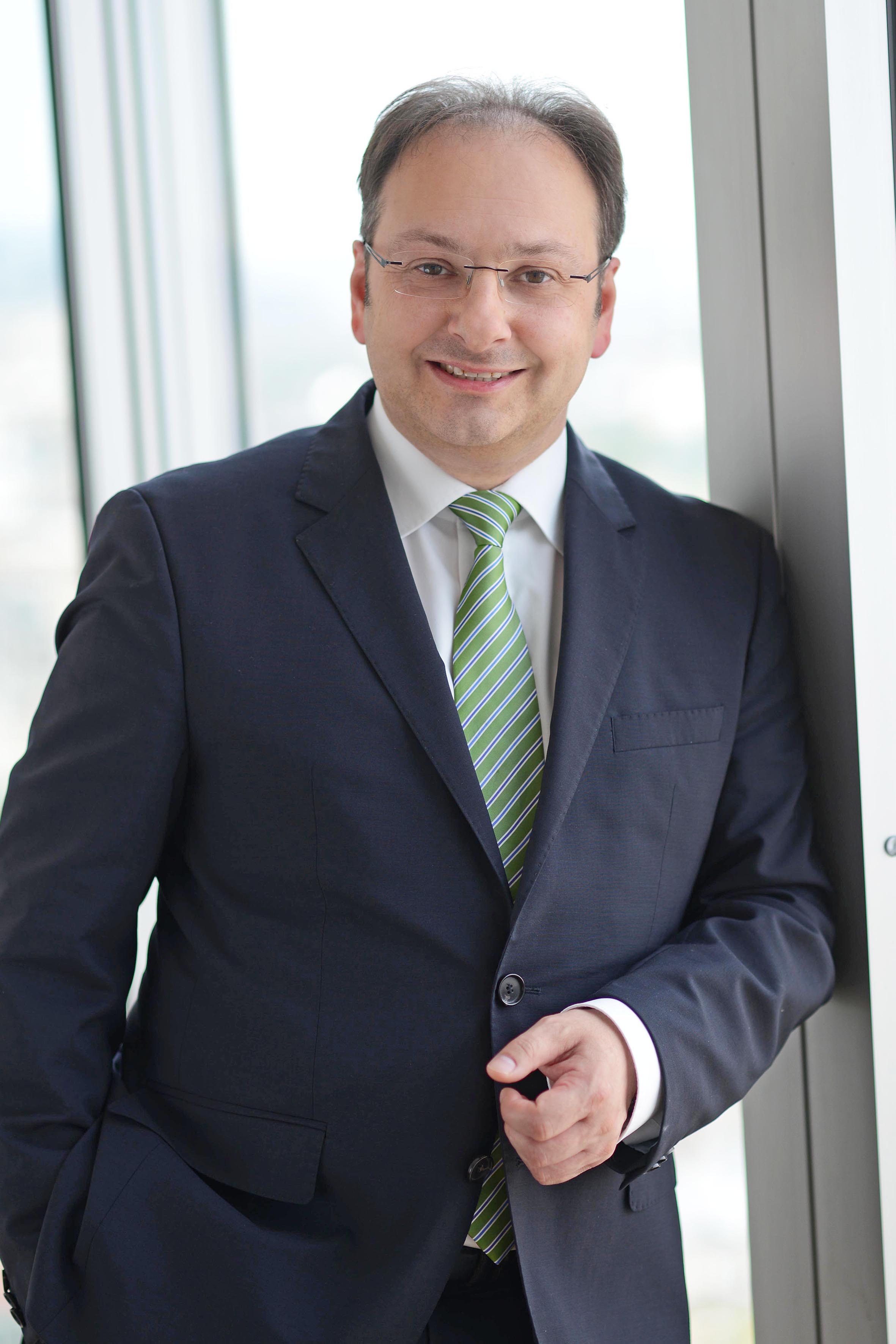 A man dressed in a dark suit with a white shirt and a green striped tie smiles while leaning against a window. The background shows a bright, blurred cityscape outside the glass. His demeanor is professional and approachable.