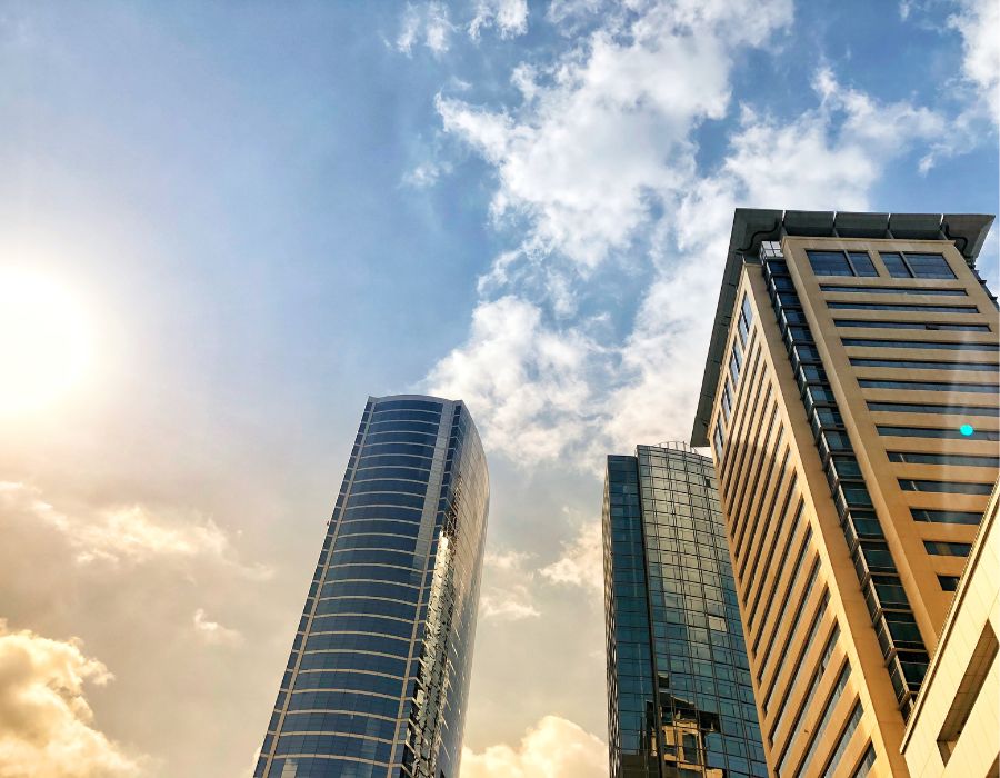 A view of modern skyscrapers reaching towards a bright sky with scattered clouds. The buildings feature glass facades, reflecting sunlight and displaying various architectural styles. The scene captures a vibrant urban atmosphere.