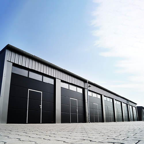 A modern storage facility featuring multiple large black doors, set against a clear blue sky. The building has a sleek, industrial design with a metallic roof and a paved foreground.
