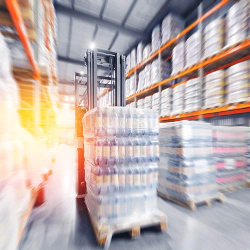 A forklift is lifting a pallet of bottled goods in a warehouse. Shelves stocked with various items are visible in the background, creating a busy storage environment. The lighting suggests an active work setting.