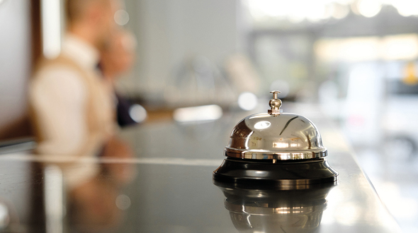 A shiny silver service bell sits on a polished counter, with blurred figures of hotel staff in the background. The setting conveys a welcoming atmosphere, typical of a reception area in a hotel.