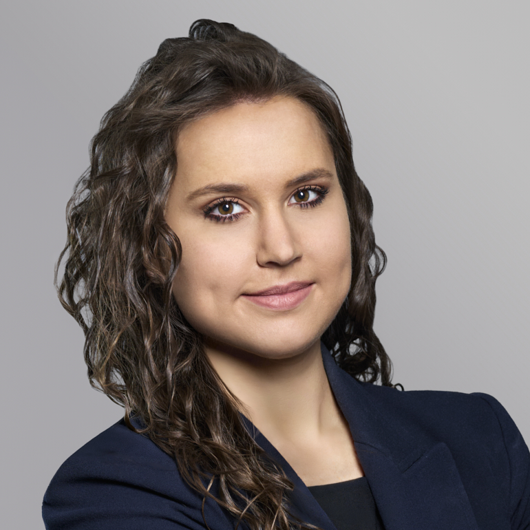 Porträt einer Frau mit lockigem, braunem Haar, die lächelt und in ein schwarzes Blazer-Jackett gekleidet ist. Sie schaut direkt in die Kamera, vor einem hellgrauen Hintergrund.