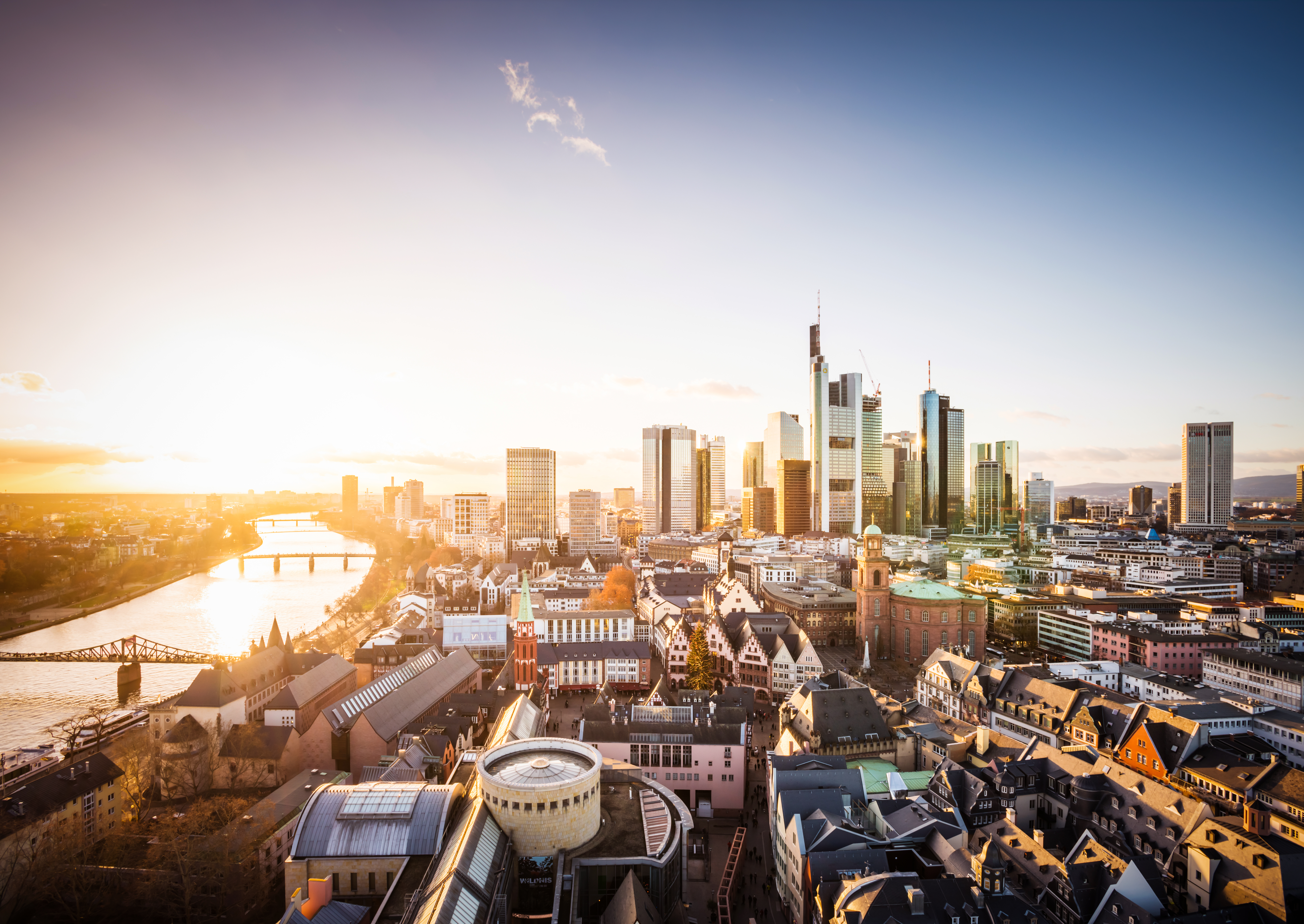 A panoramic view of a city skyline during sunset, showcasing modern skyscrapers alongside traditional buildings. A river flows through the scene, with bridges connecting both sides. The warm glow of the setting sun reflects off the glass facades, creating a vibrant atmosphere.