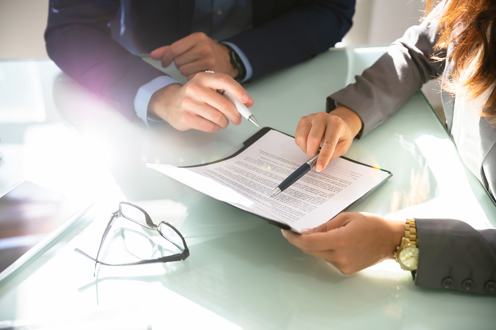 A close-up of two hands engaged in a discussion over a document on a table. One hand points to the text on a clipboard, while the other holds a pen, indicating a collaborative review or signing process.