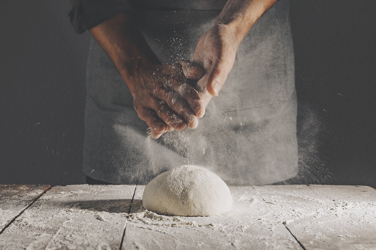 A pair of hands is shown kneading dough on a wooden surface dusted with flour. The hands are pressing down and creating a cloud of flour, highlighting the process of baking or cooking. The background is dark, focusing attention on the action of preparing the dough.