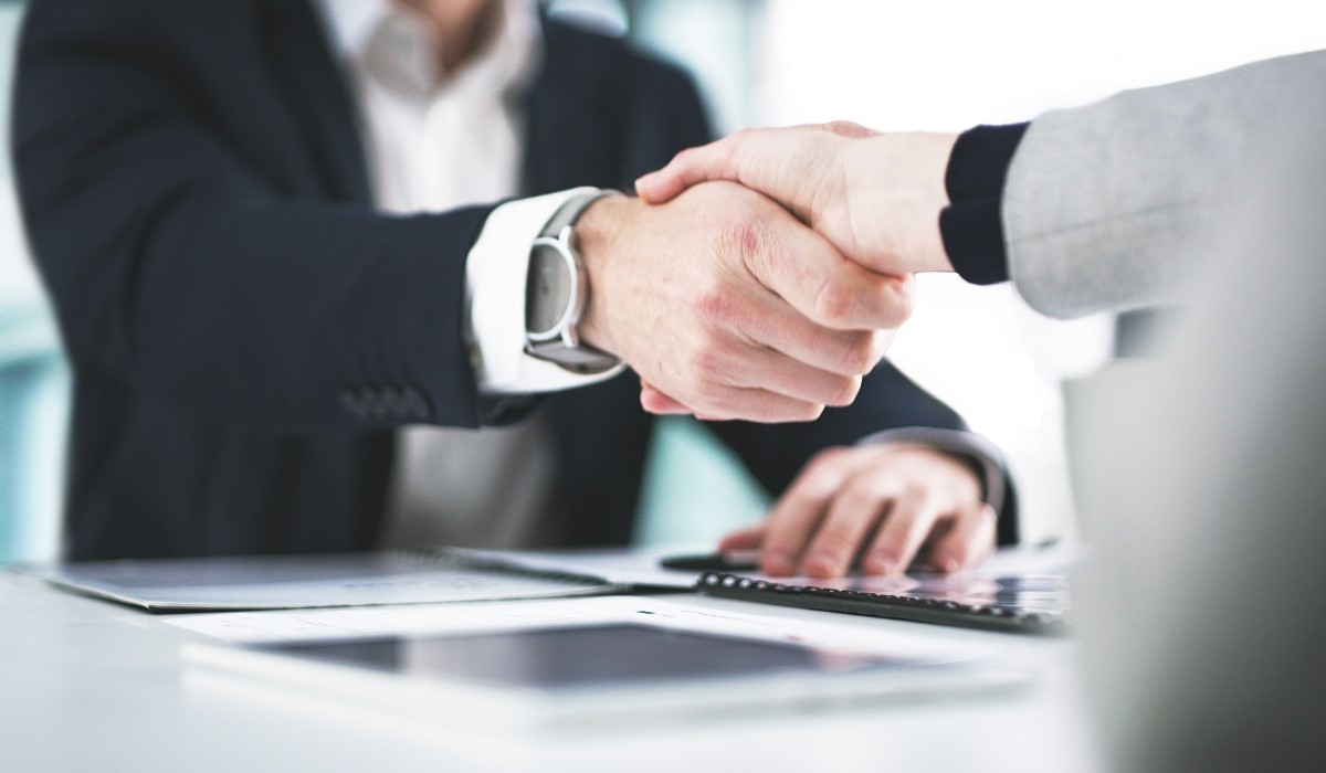 Two individuals are shaking hands in a business setting, signifying a partnership or agreement. A table with documents and electronic devices is visible in the foreground, emphasizing a professional atmosphere.