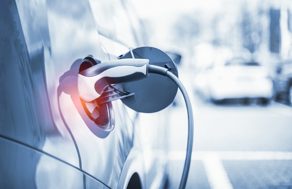 A close-up of an electric vehicle's charging port with a charging cable connected. The background is blurred, suggesting a parking area. The focus highlights the connection point, emphasizing the vehicle's electric charging process.