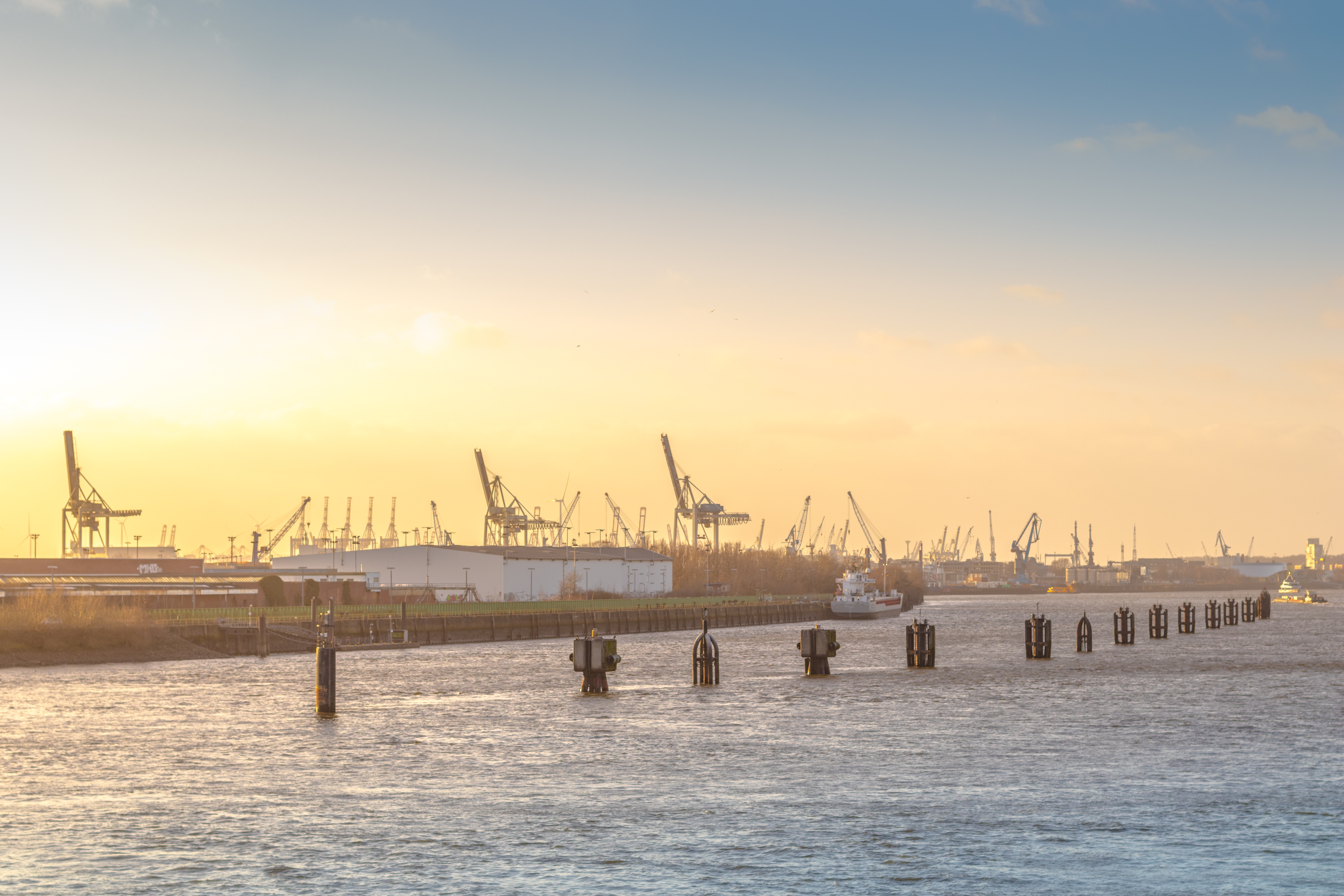 A river scene at sunset shows industrial cranes in the background and several boats on the water. In the foreground, wooden posts rise above the surface of the river, creating a structured pattern along the shoreline. The atmosphere is calm and serene.