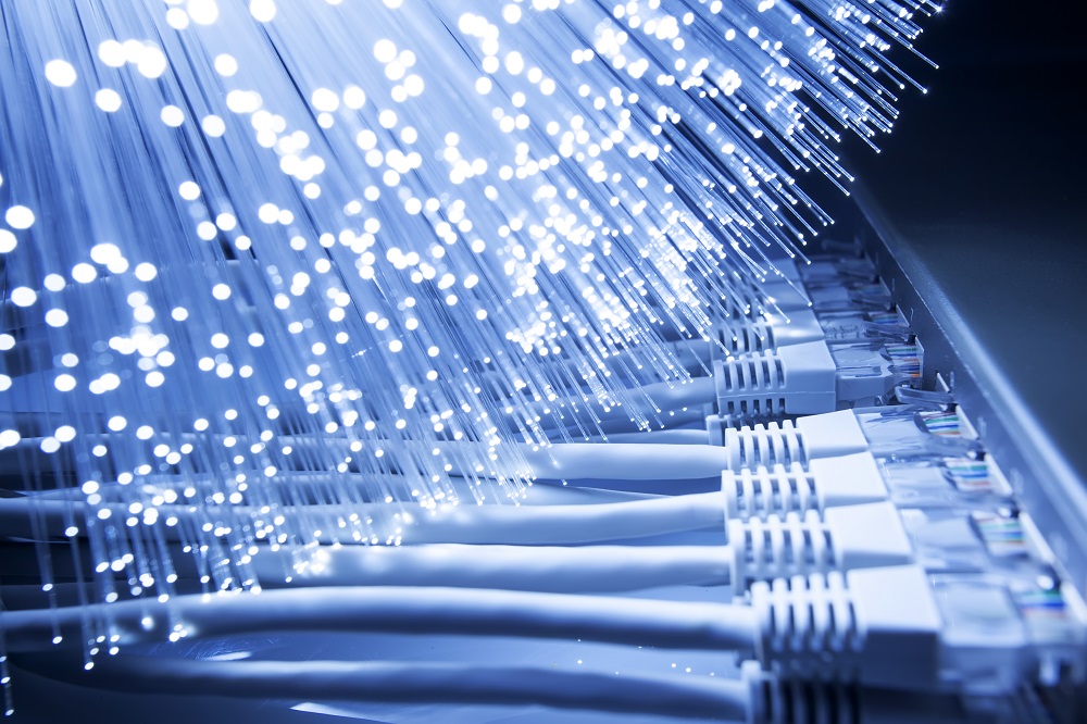 Close-up of fiber optic cables illuminated by light, showcasing their glowing strands. Below, several connected Ethernet cables are visible, indicating a network setup. The image emphasizes the technology behind high-speed internet connectivity.