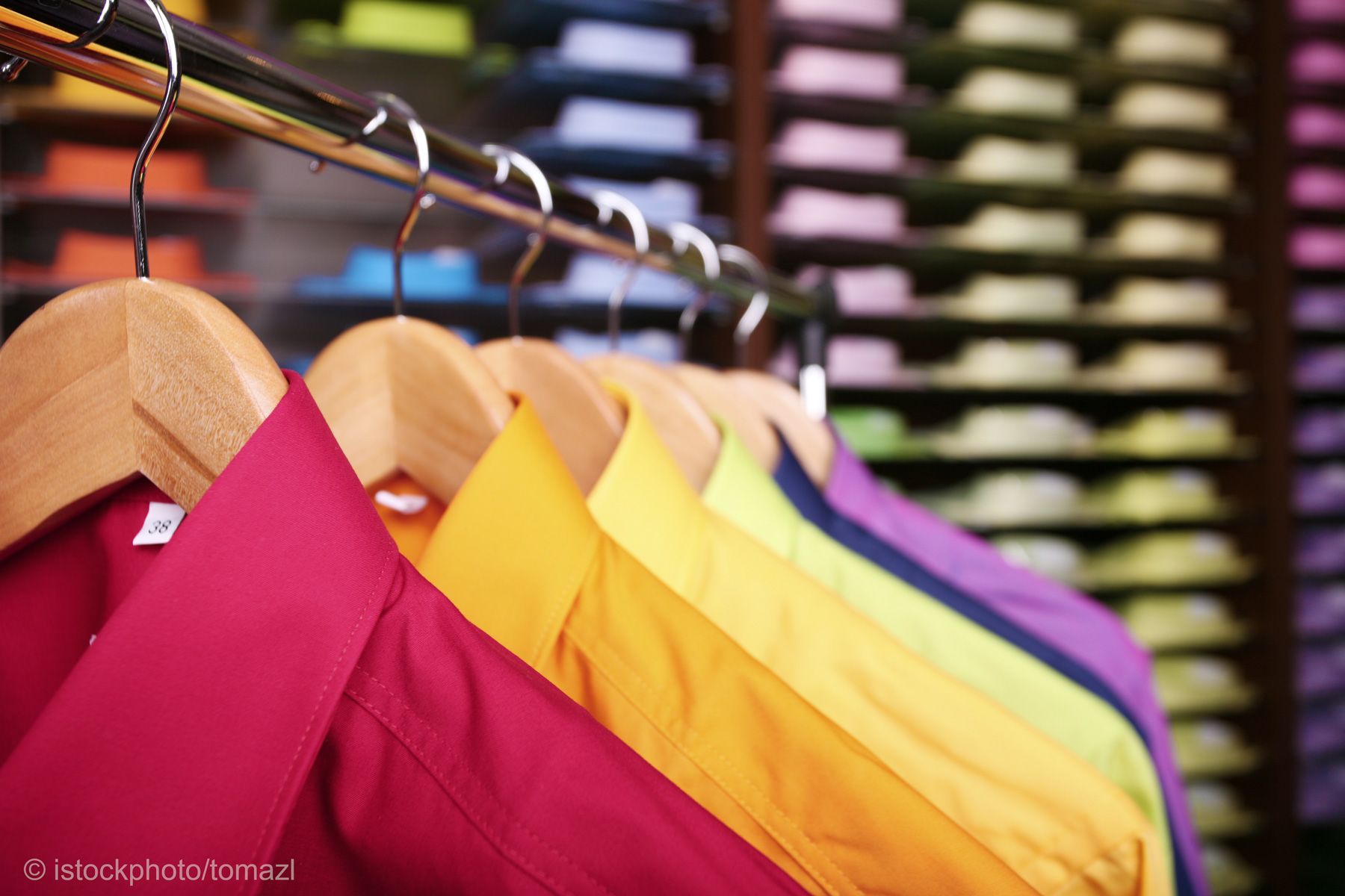 A row of colorful shirts, including red, yellow, and purple, hanging on wooden hangers. In the background, neatly stacked papers in various colors are visible, creating a vibrant and organized display.