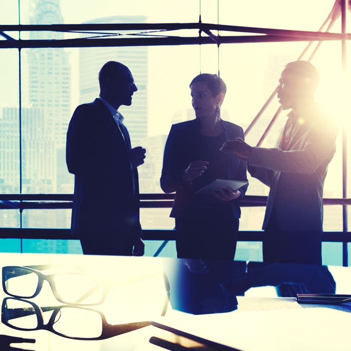Three professionals engage in a discussion in a well-lit office with large windows showcasing a cityscape. One person holds a tablet, while the others appear to be sharing ideas. A pair of glasses is placed in the foreground, emphasizing a scholarly or business context.