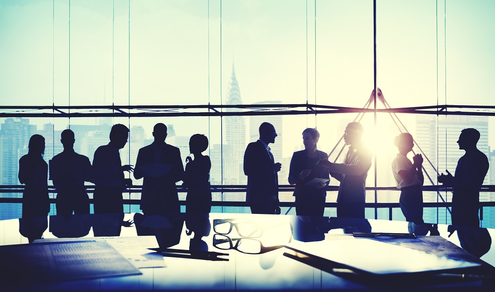 Silhouettes of business professionals engage in conversation near a large window with a city skyline in the background. The sun is setting, creating a bright glow behind them. On a table in the foreground, there are glasses and scattered documents.