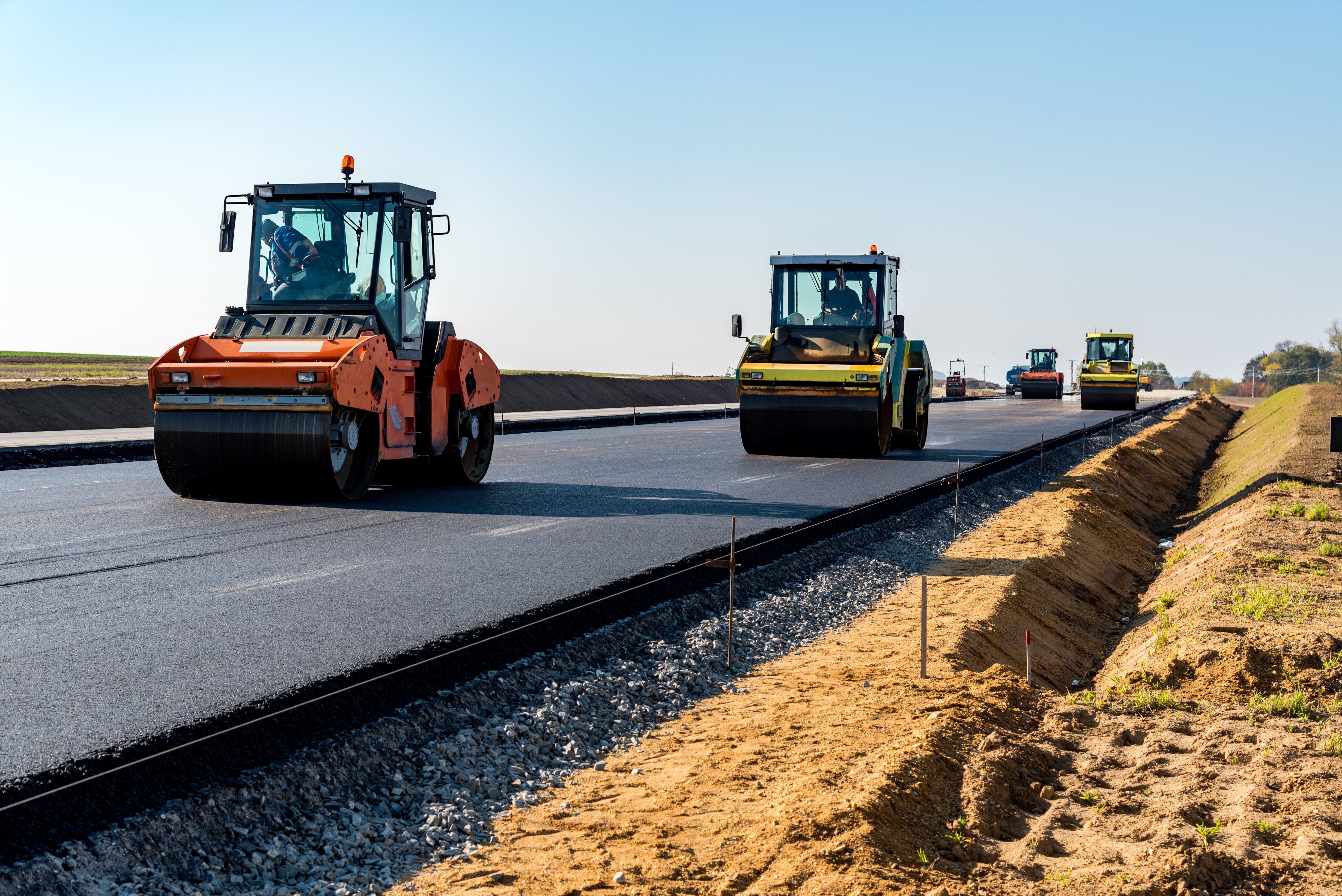 A construction site with multiple heavy-duty road roller machines working on a newly paved road. The scene shows various rollers engaged in compaction, with an open, blue sky in the background. The gravel and dirt along the road’s edge are visible, indicating ongoing construction.