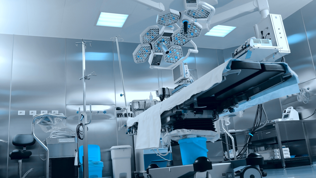 A modern surgical room featuring an operating table at the center, illuminated by overhead surgical lights. Medical equipment is arranged around the room, including a vital signs monitor and an IV stand, all set against a stainless steel backdrop.