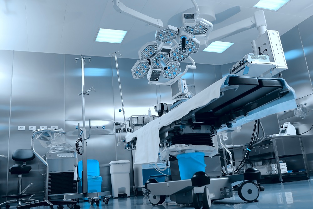 A modern surgical room featuring an adjustable operating table with a sterile sheet, overhead surgical lights with multiple fixtures, and medical equipment including an IV stand and monitors. The space has metallic walls, emphasizing a clean and clinical environment.