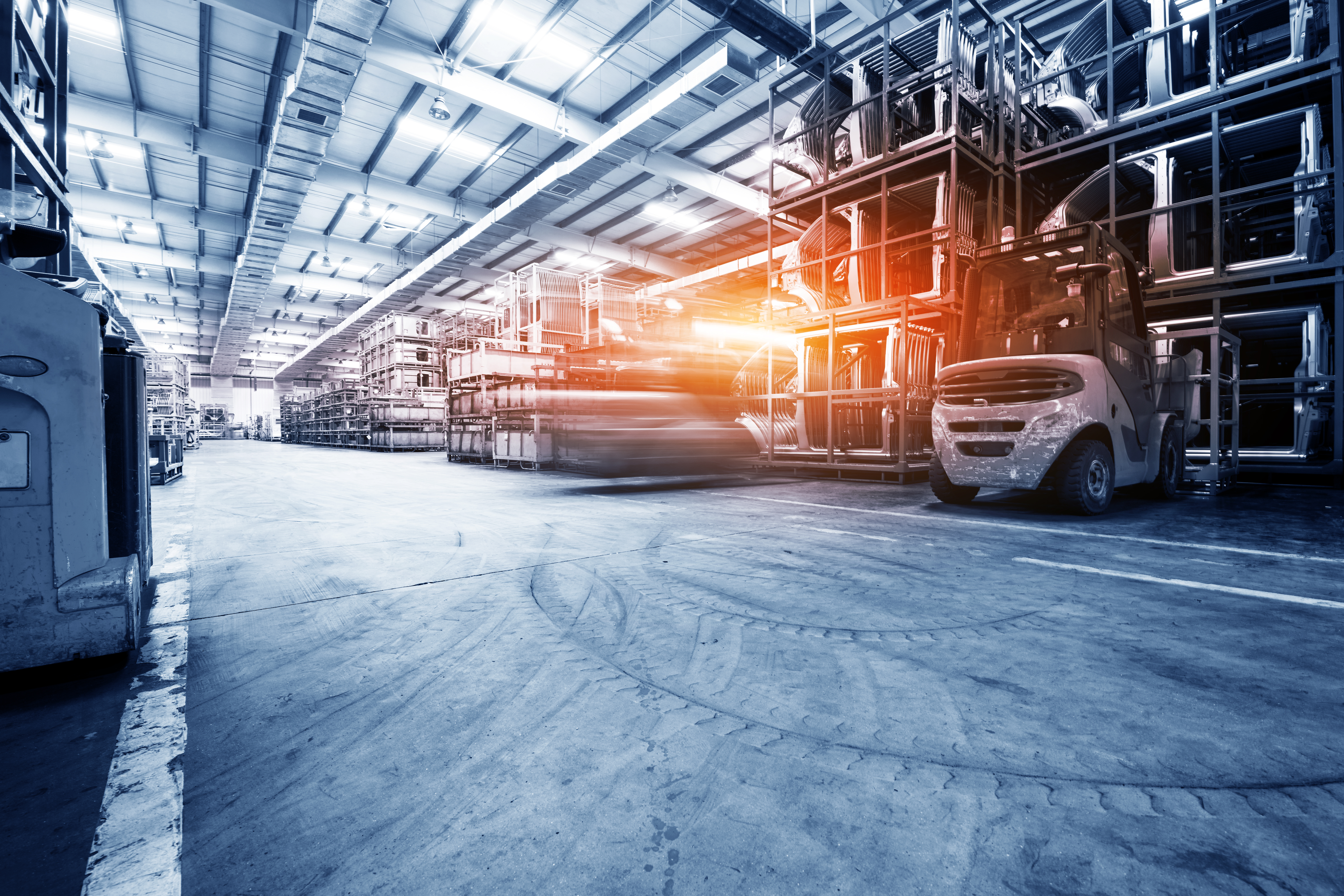 A large industrial warehouse interior with high ceilings is filled with stacked pallets and shelves. In the foreground, a forklift is parked, while another forklift is seen in motion, emphasizing a busy work environment. Bright overhead lights illuminate the space.