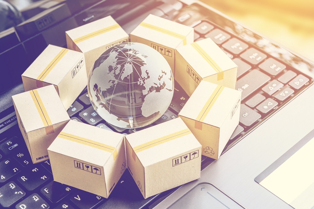A clear glass globe depicting a world map is surrounded by several small cardboard boxes with yellow stripes. The scene is set on a laptop keyboard, symbolizing global shipping and e-commerce.