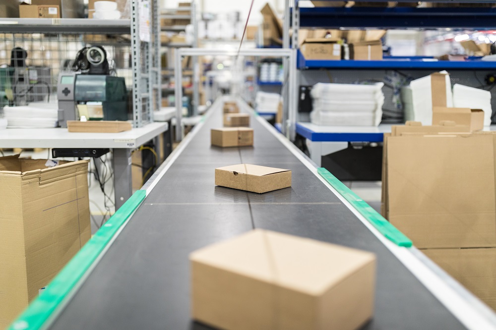A conveyor belt lined with several small, brown cardboard boxes is showcased in a warehouse setting, surrounded by shelves filled with packaging materials. The scene conveys a sense of organized shipping or sorting activity.