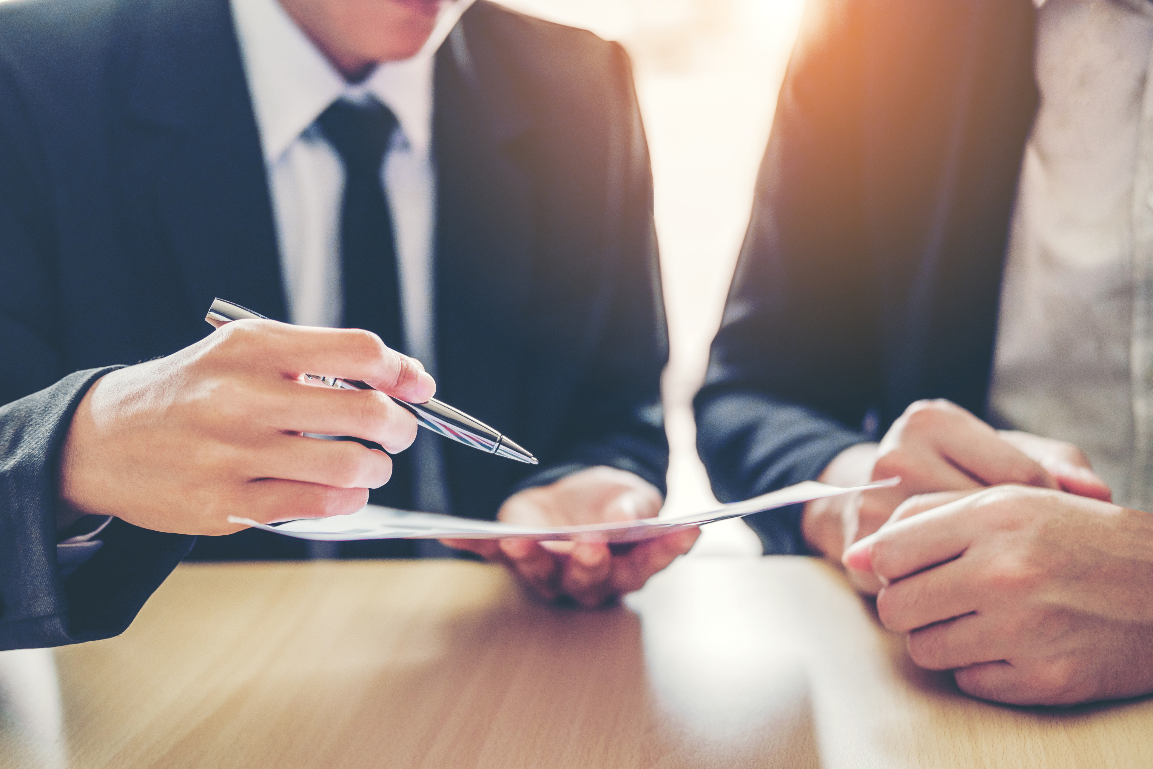 Two individuals in business attire are engaged in a discussion. One holds a pen and is pointing to a document on the table, while the other listens attentively, resting their hands on the table. The setting suggests a professional meeting or negotiation.