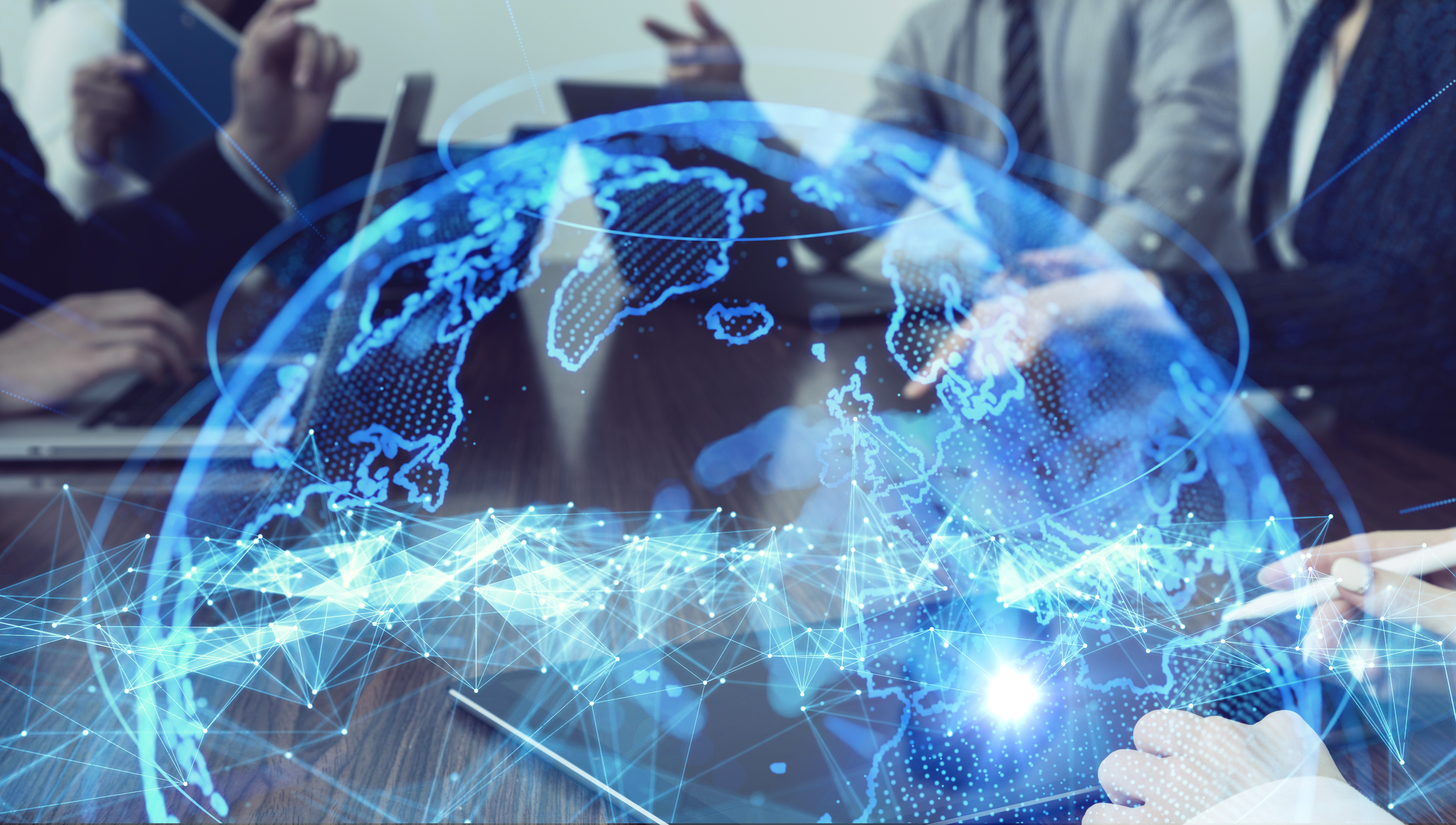 A digital globe with a network of lines and dots overlays a meeting scene. People are engaged in discussion, using laptops and tablets. The focus is on collaboration and technology, symbolizing global communication and connectivity in a professional environment.