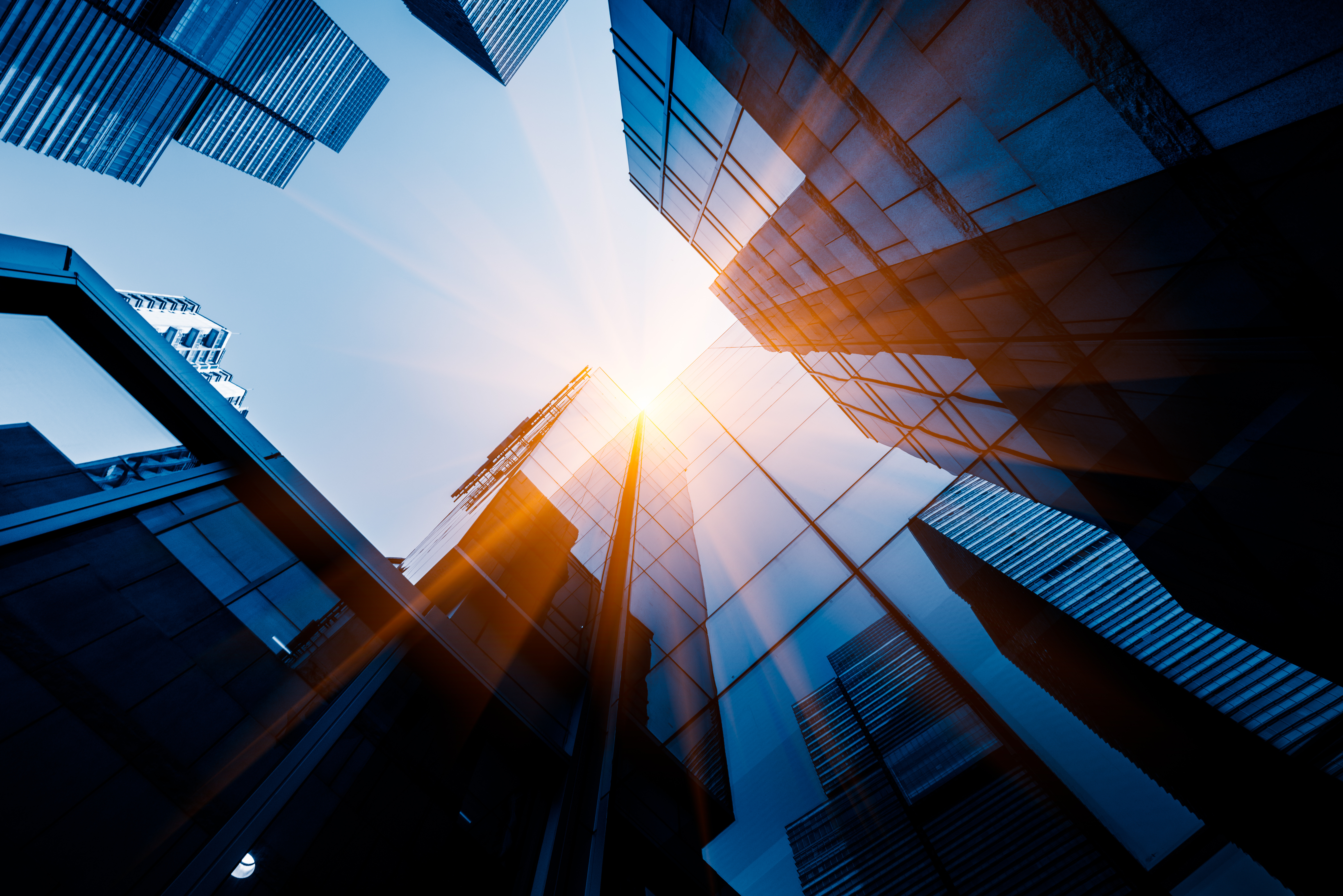 A low-angle view of modern glass skyscrapers reflecting sunlight against a blue sky. The image captures beams of light breaking through the structures, creating a vibrant atmosphere of urban elegance and energy.