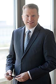 A professional man in a suit stands by a window, smiling and holding a mobile device. The background features soft lighting and a blurred outdoor view, suggesting an office environment.