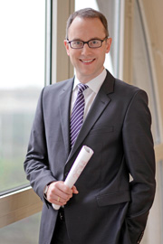 A professionally dressed man with glasses stands by a large window, smiling confidently. He holds a rolled document in one hand, suggesting a business or professional setting. The background is softly blurred, emphasizing his presence and demeanor.