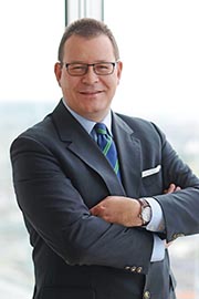 A professionally dressed man stands with his arms crossed, smiling confidently. He wears glasses and a dark suit with a green and blue striped tie. The background suggests an office setting with a view of the city skyline.