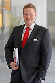 A man in a dark suit with a red tie stands confidently, holding a folder. He is in a professional setting, suggesting a business context. His expression is friendly and approachable.